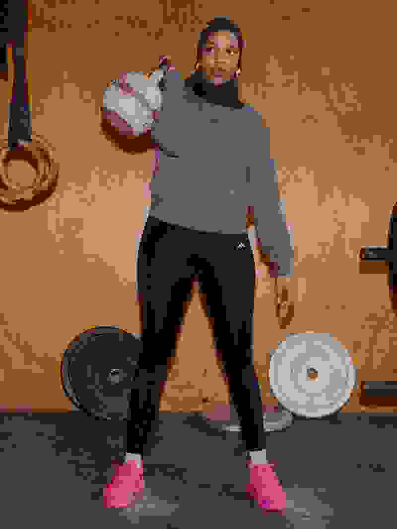 A person wearing a gray sweatshirt and black leggings stands in a gym setting, holding a weight in their hands, with various exercise equipment visible in the background.