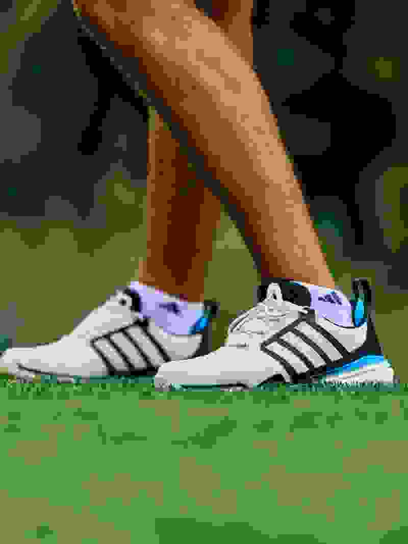 The image shows a pair of white and black athletic shoes with blue accents, resting on a grassy surface against a blurred green background.