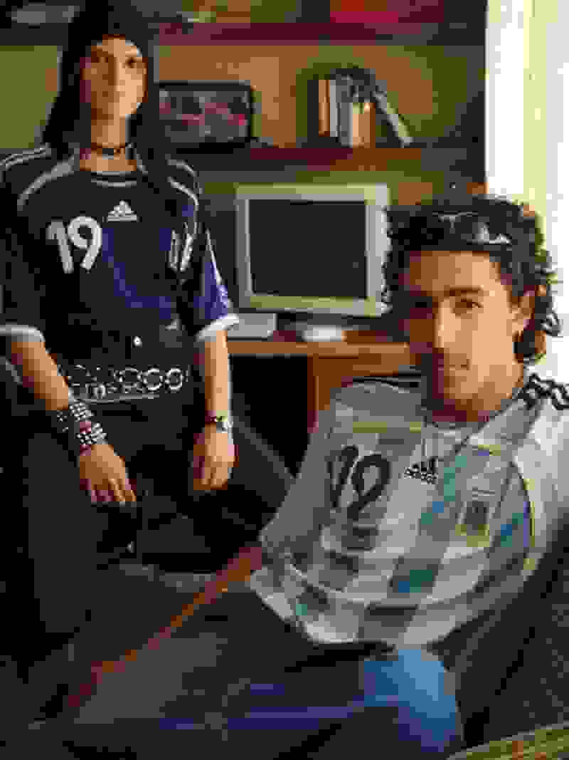 Two individuals, a man and a woman, are seated in a room with shelves and a television visible in the background. They are both wearing jerseys with the number 19 on them.