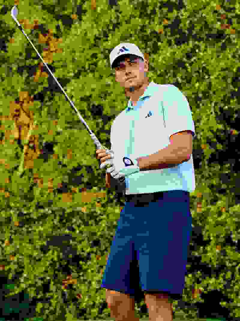 A man in a light blue polo shirt and white cap is holding a golf club and standing in a lush, green outdoor setting.