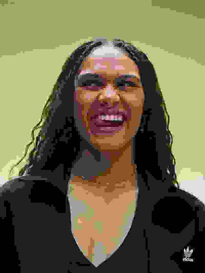 A smiling young woman with curly dark hair wearing a black top stands against a vibrant green background.