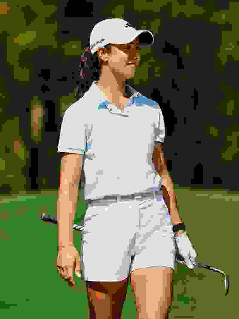 A young woman in a light blue polo shirt and white shorts stands on a grassy golf course, holding a golf club and wearing a baseball cap.