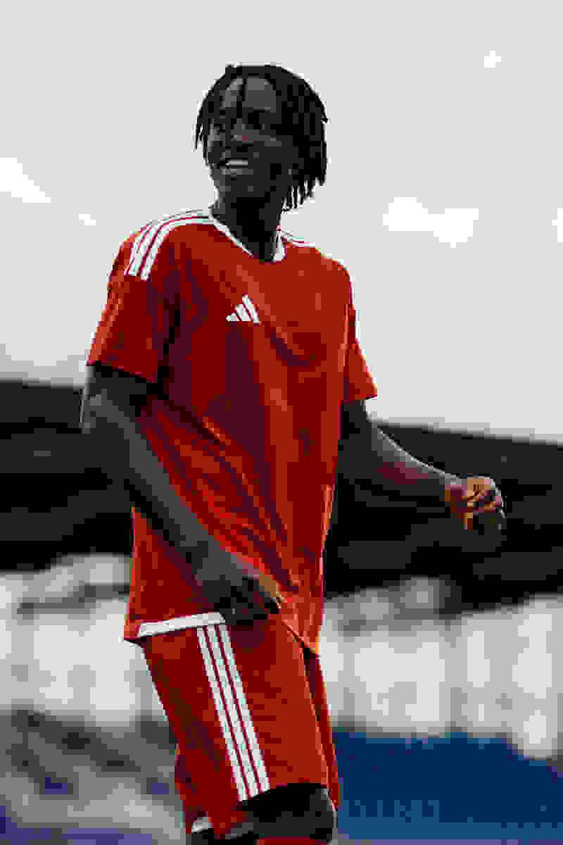 A smiling young man wearing a red Adidas jersey stands in front of a body of water with mountains in the background.
