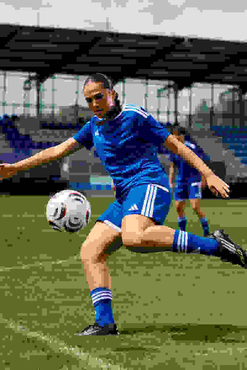 A female soccer player in a blue uniform is running on a grassy field, with a stadium-like structure visible in the background.