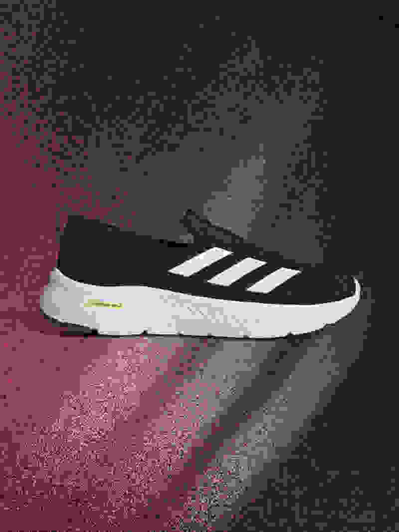 A black and white athletic shoe with the Adidas logo sits on a red and black textured background, casting a subtle shadow.