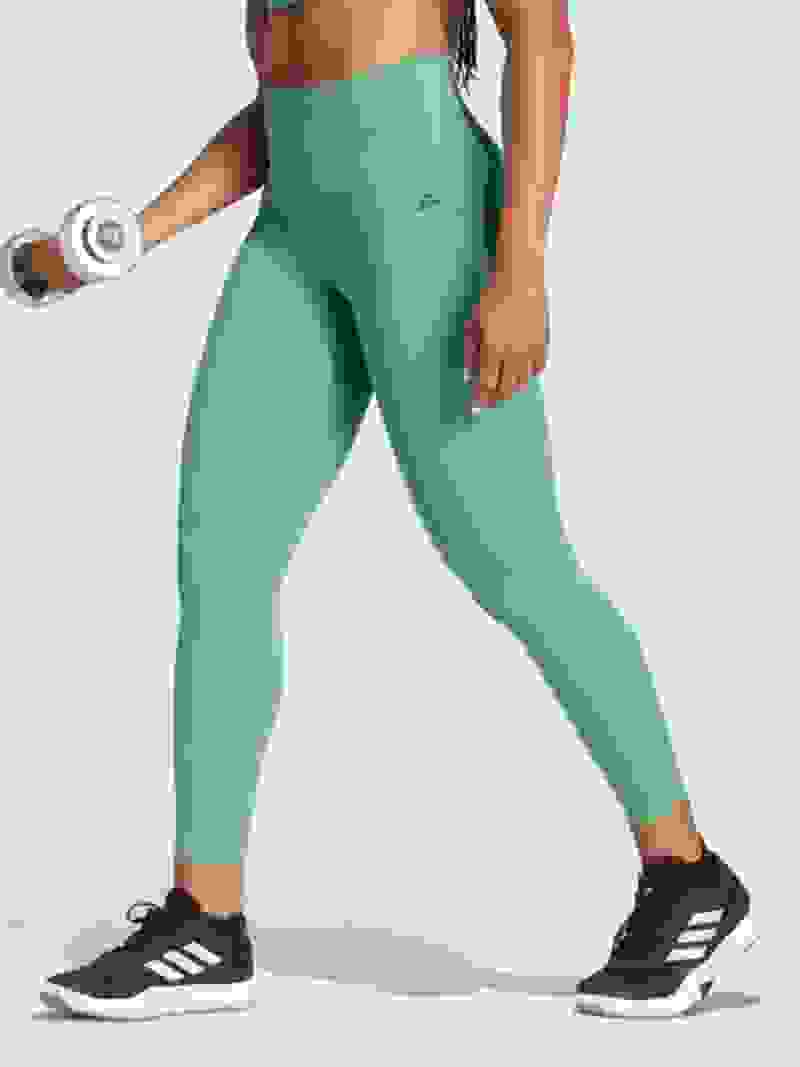 De afbeelding toont een persoon die een groene sportlegging en zwarte sneakers draagt en in een studio staat met een achtergrond van wit.