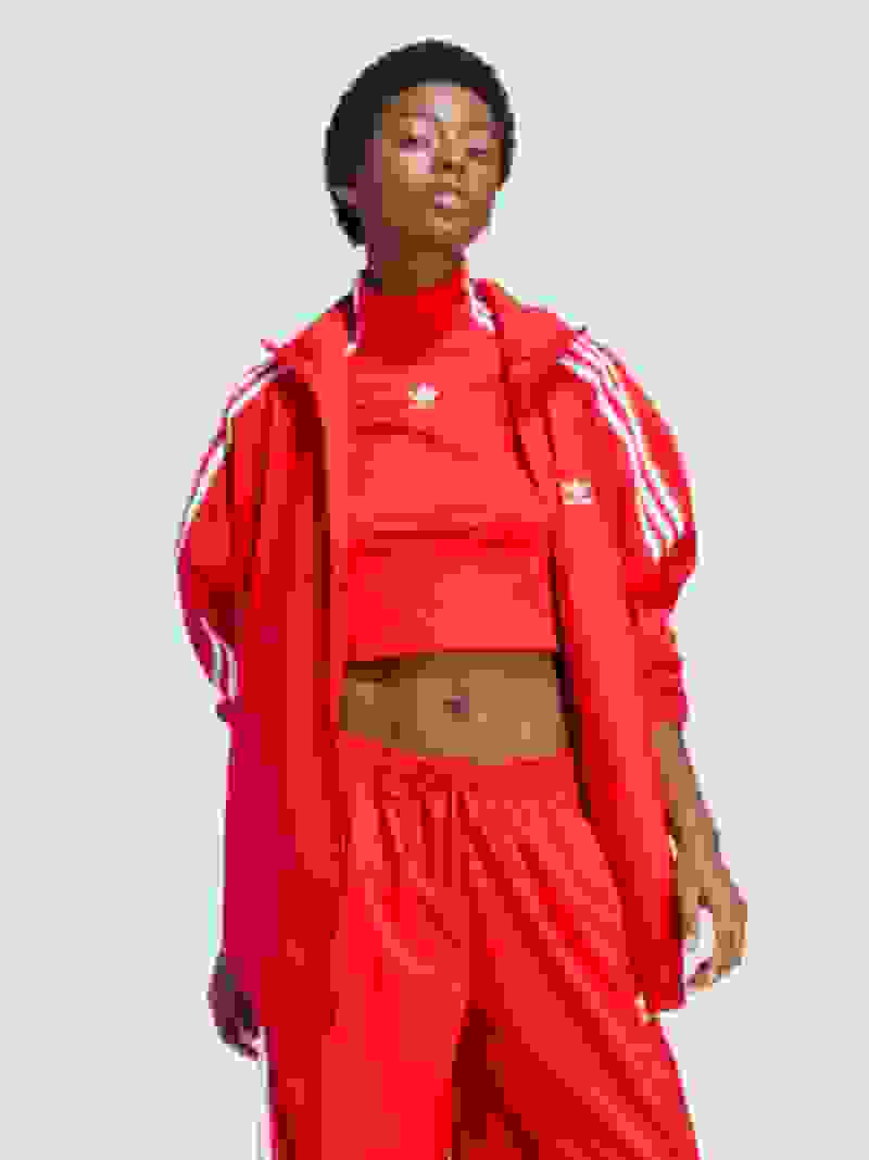 Een jonge vrouw in een felrood adidas trainingspak staat op een achtergrond in effen wit, haar zelfverzekerde en gefocuste expressie.