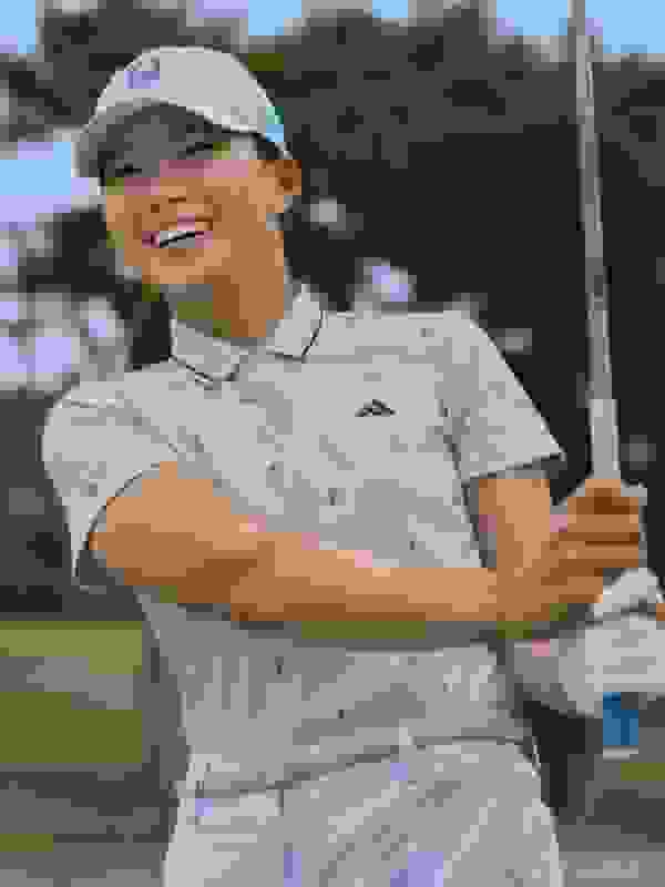 A young woman in a golf outfit is smiling and holding a golf club on a grassy golf course with trees in the background.