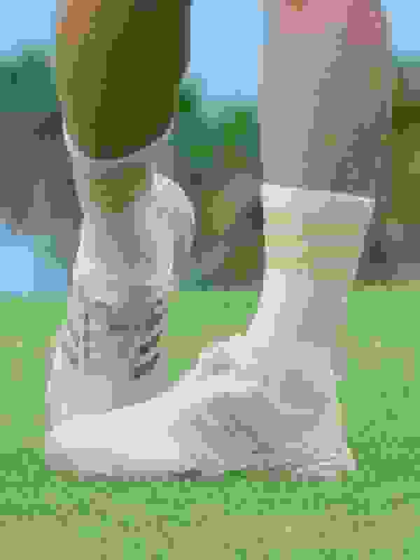 The image shows a person's lower legs and feet wearing white athletic shoes with yellow stripes on a grassy field.