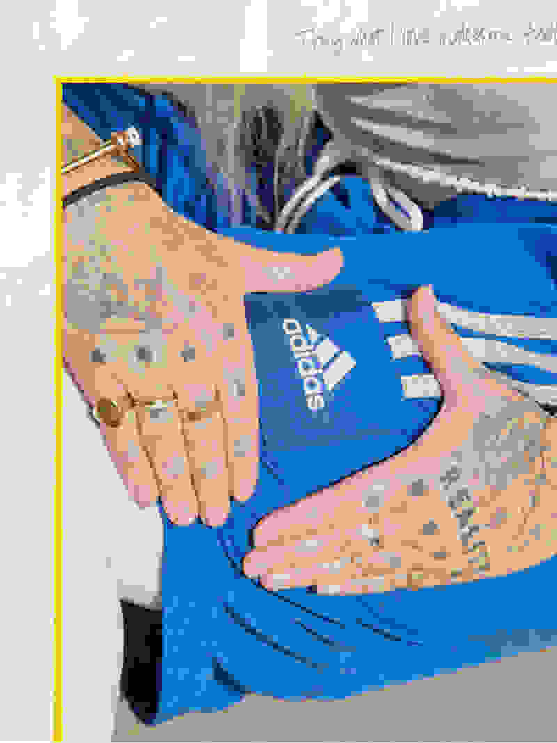 The image shows a person's hands holding an Adidas-branded blue clothing item, with visible tattoos on the person's hands and arms.