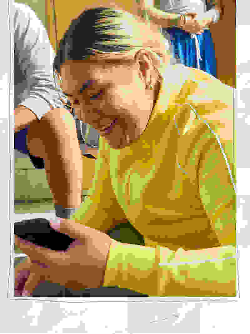 A young woman in a bright yellow sweater is smiling and using a smartphone while sitting in a colorful, cluttered room.