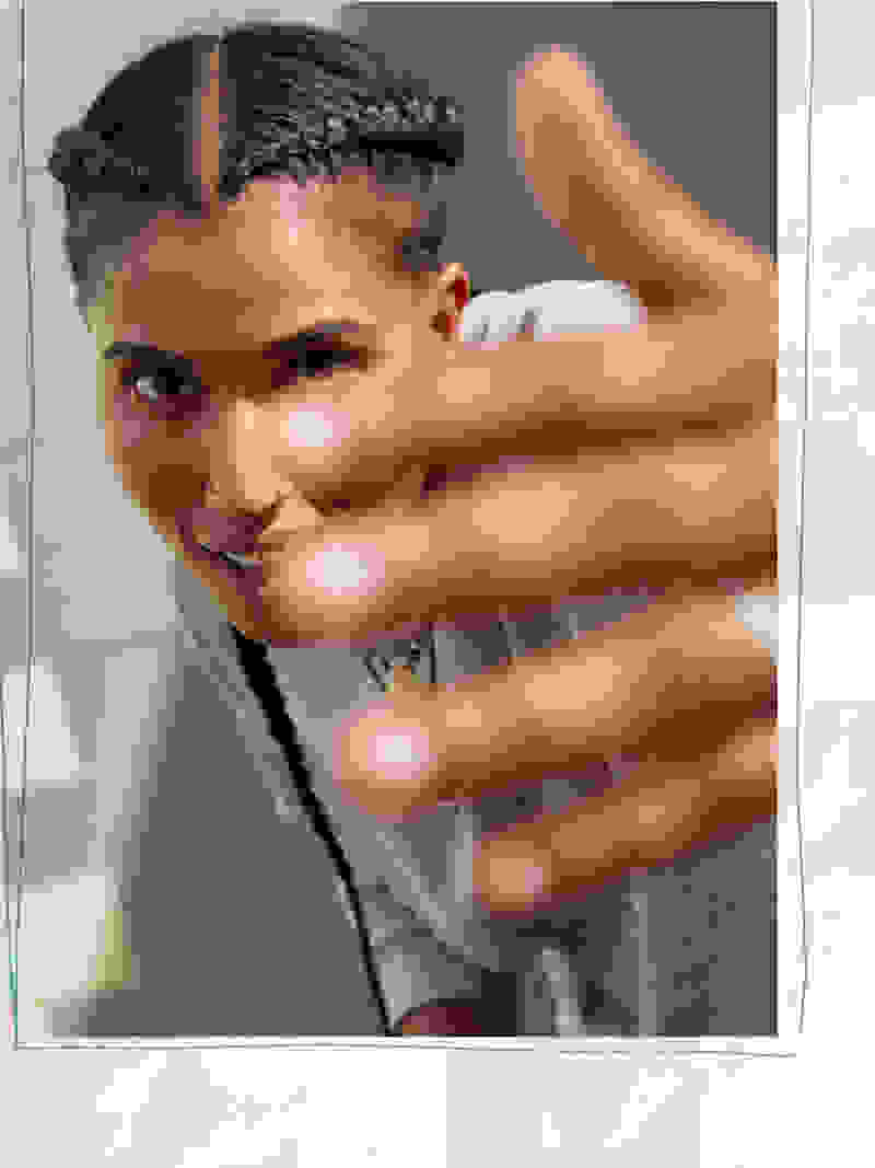 The image shows a person with braided hair making a hand gesture in the foreground, with a blurred background.
