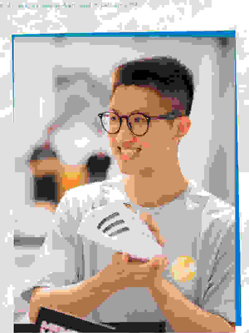 A young man wearing glasses and a white t-shirt is smiling and holding a pair of white sneakers in the foreground, with a blurred background of what appears to be an indoor setting.