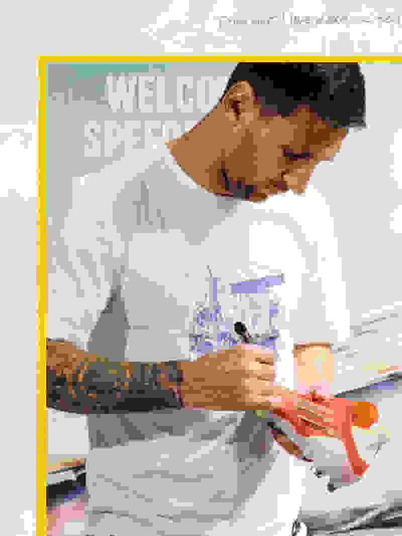 A man wearing a white t-shirt with a graphic design is holding an orange toy gun in the foreground, with a "Welcome Speed" sign visible in the background.