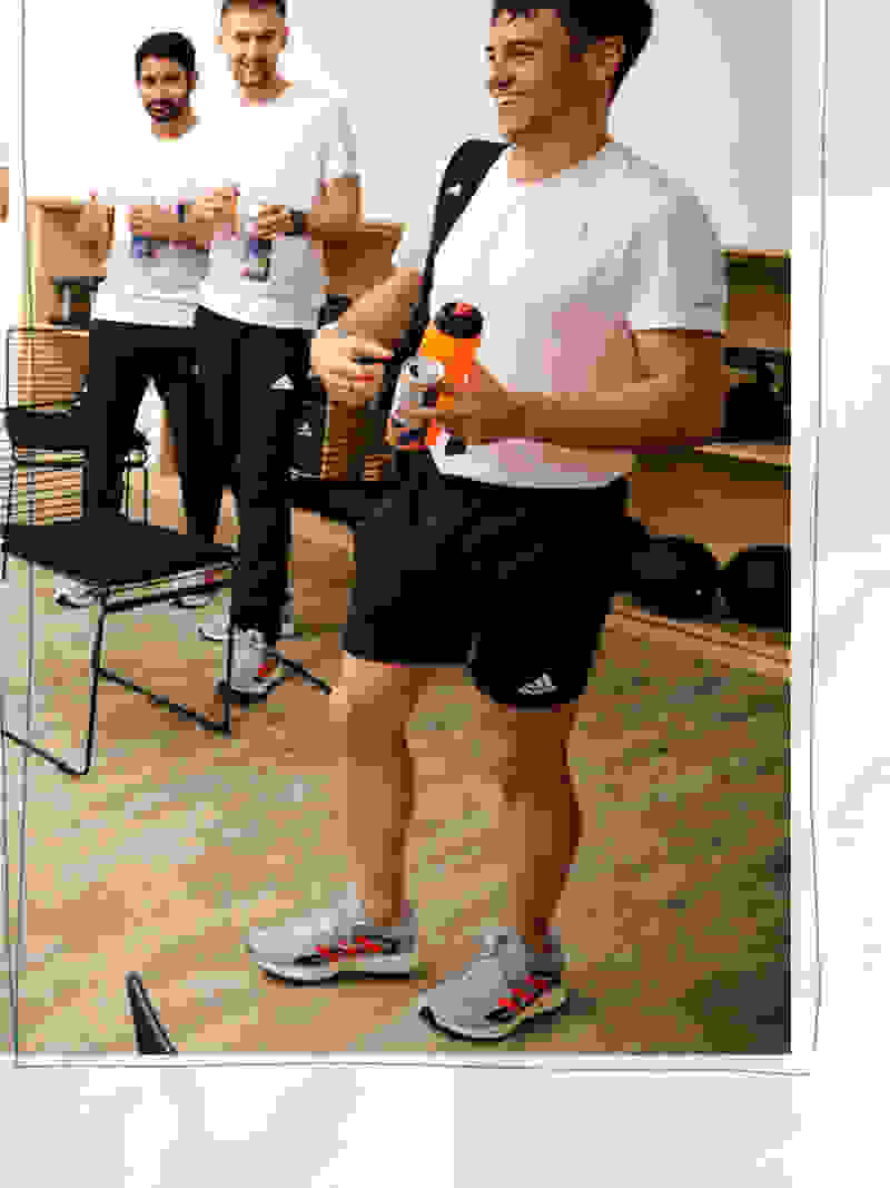 The image shows three men in a fitness studio, with one man in the foreground holding a water bottle and wearing athletic clothing.