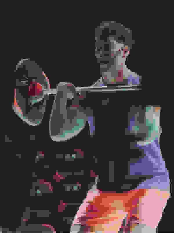 A muscular man in a blue shirt and orange shorts is lifting weights in a dimly lit gym setting.