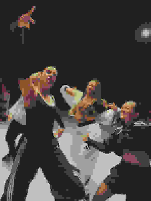 The image depicts a group of people engaged in a lively dance performance, with the foreground featuring a woman in a black outfit striking a dynamic pose while the others around her appear to be moving in synchronization.