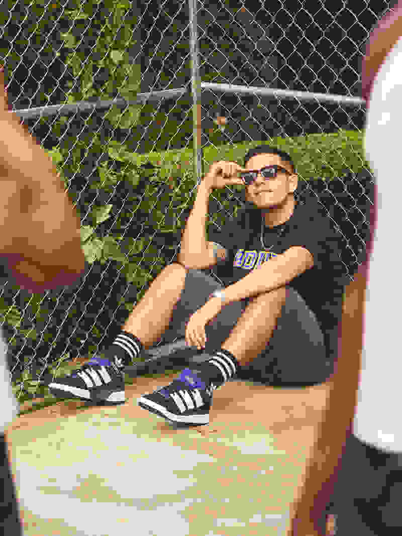 A person wearing a black shirt, sunglasses, and sneakers is sitting on the ground in front of a chain-link fence with greenery in the background.