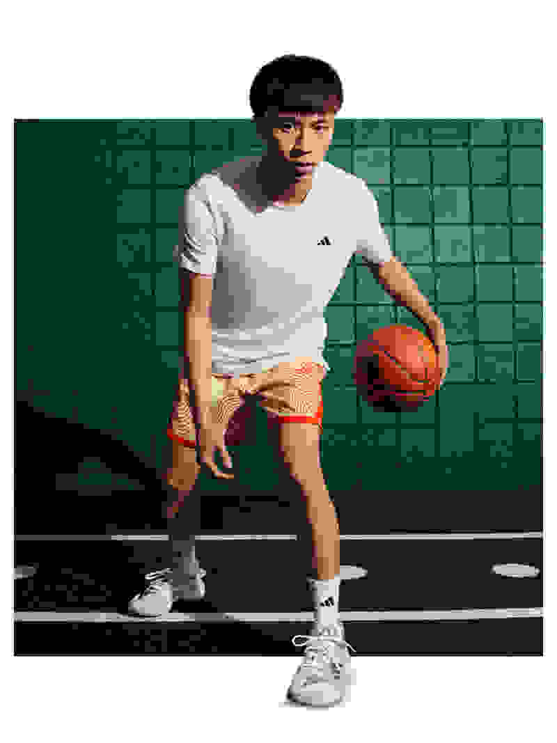 A young man in a white t-shirt and orange shorts stands on a basketball court, holding an orange basketball.