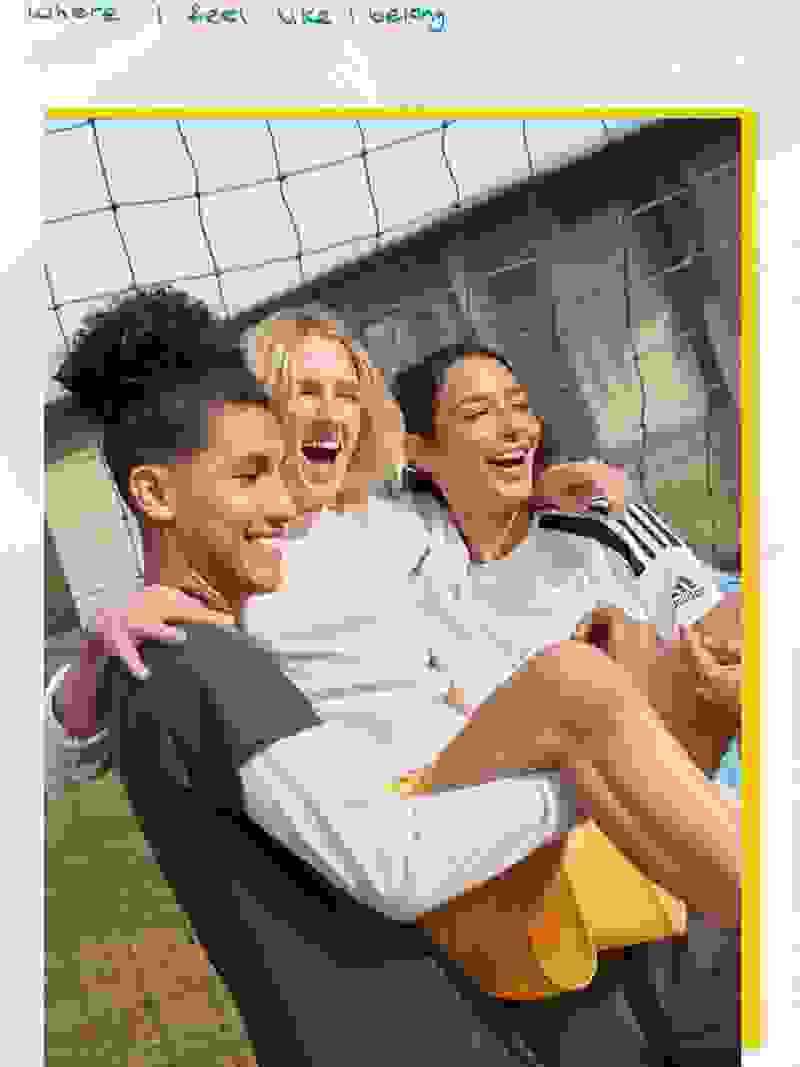 Drei junge Leute, zwei Frauen und ein Mann, umarmen sich und lachen gemeinsam auf einem Sportplatz mit einem Maschendrahtzaun im Hintergrund.