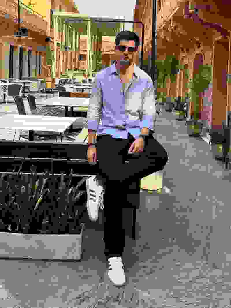 A well-dressed man sits on a bench in an outdoor setting with ornate architectural features and potted plants in the background.