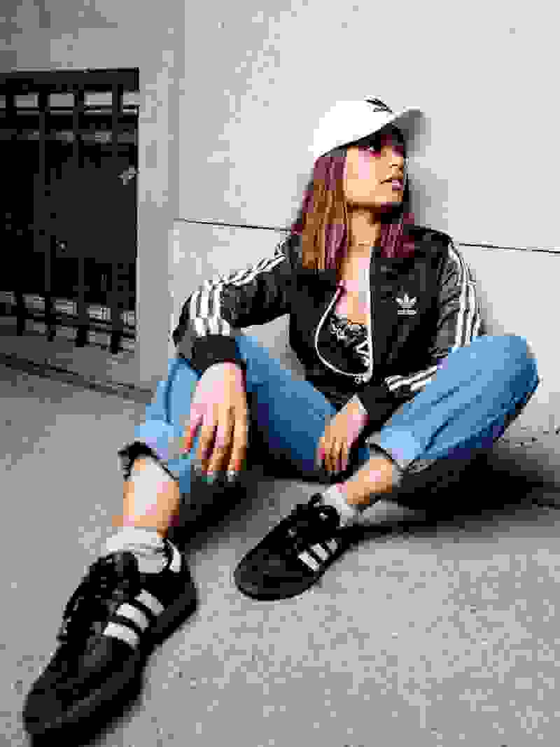 A young woman wearing an Adidas tracksuit and cap is sitting on the ground against a concrete wall.