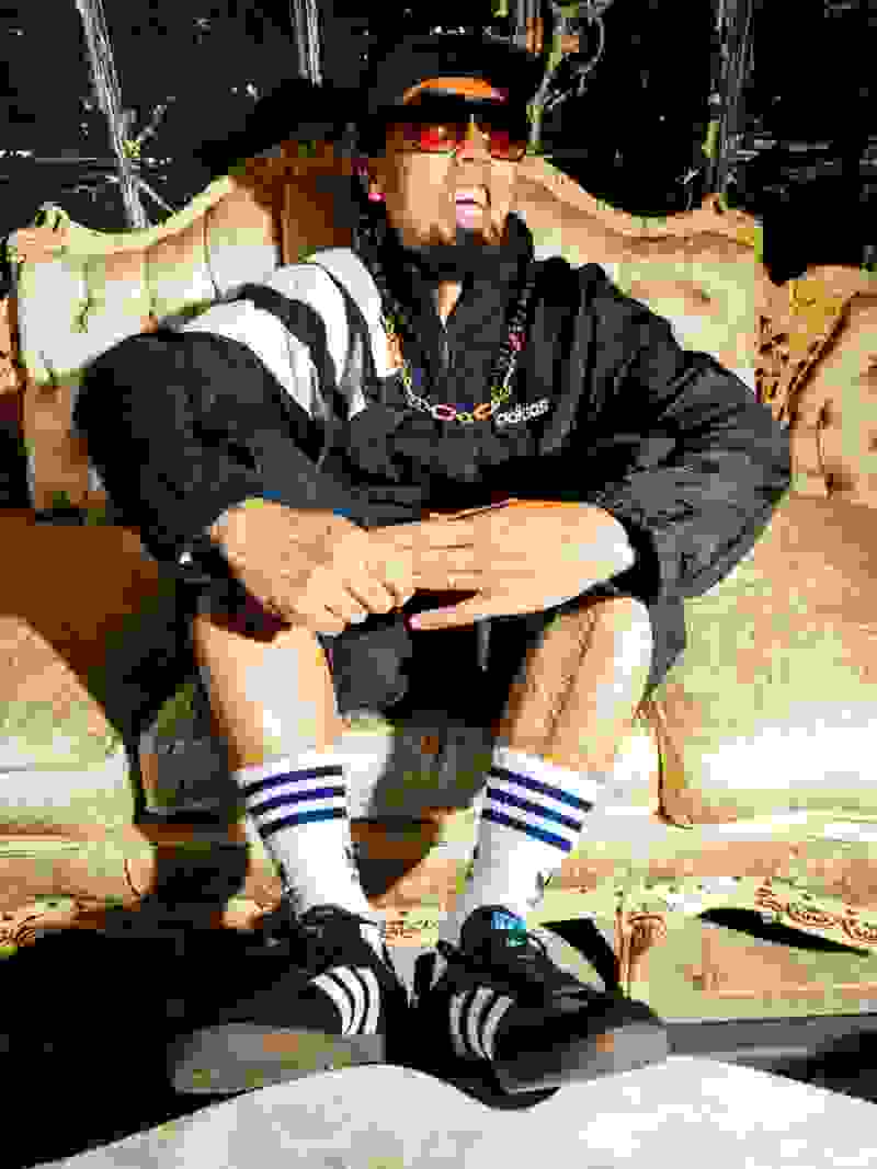 A person wearing a black jacket, hat, and sunglasses is sitting on a couch in a dimly lit room with various objects and decorations in the background.