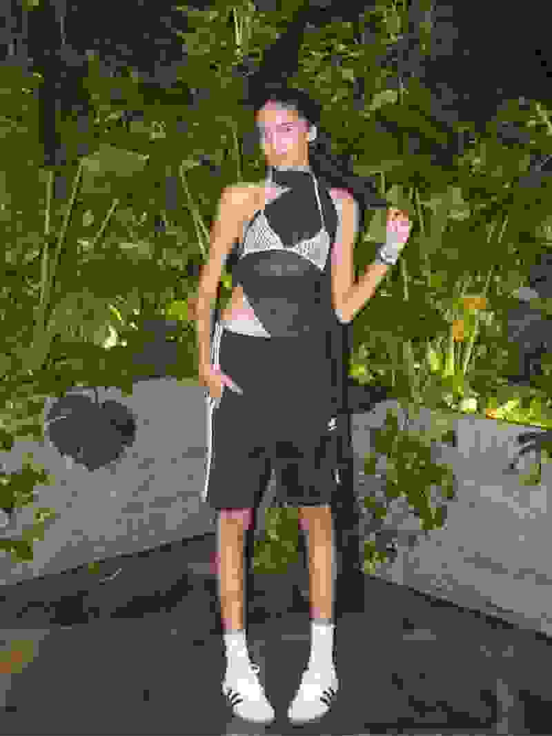 The image shows a young woman wearing a black and white outfit, standing in a lush, green outdoor setting with various plants and foliage in the background.