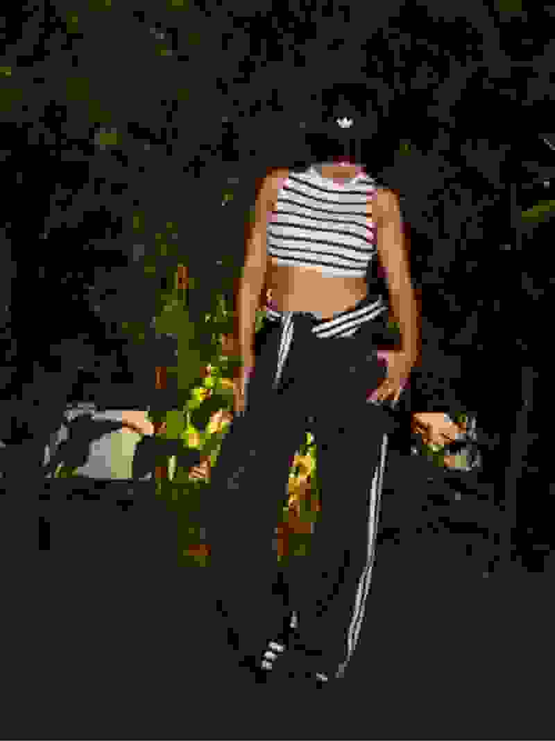 A person wearing a striped crop top and black pants stands in a lush, green outdoor setting with foliage in the background.