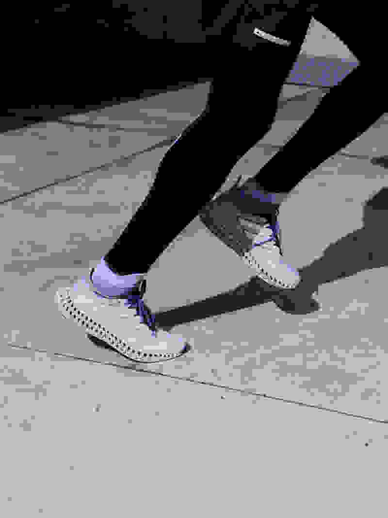 The image shows a close-up view of a white athletic shoe with a textured, patterned sole and upper. The background is dark, creating a dramatic contrast that highlights the intricate design and structure of the shoe.