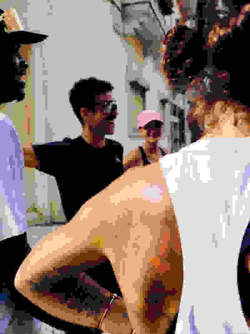 A group of people, some with tattoos, standing in an urban setting with buildings and structures visible in the background.