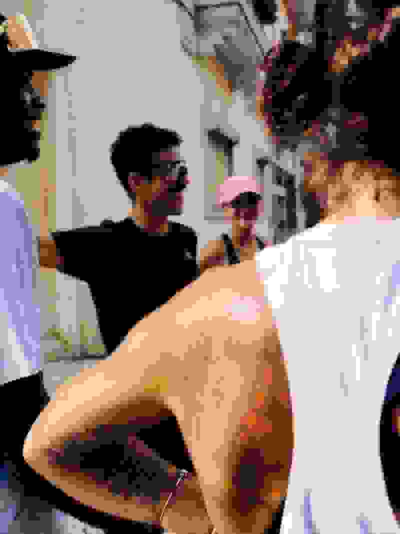 A group of people, some wearing hats and sunglasses, are gathered in an urban setting with buildings and structures visible in the background.