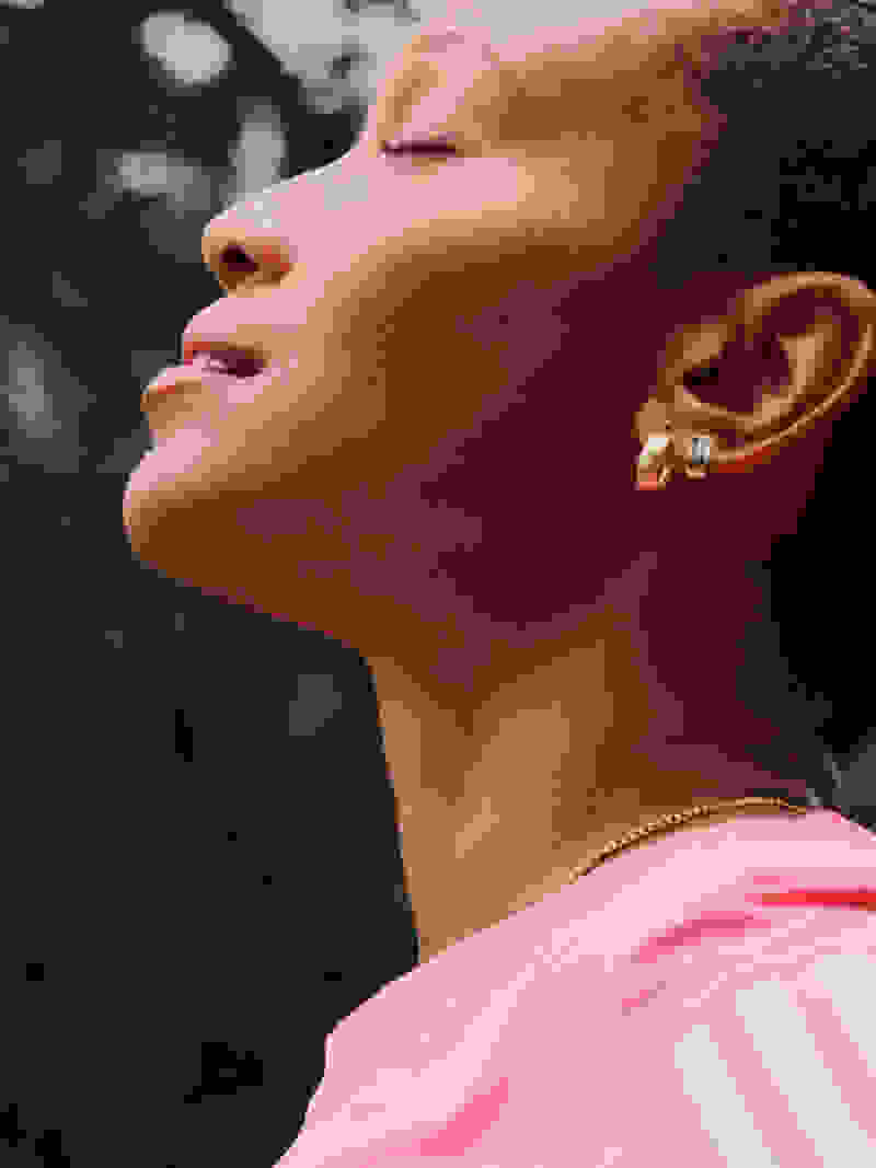 A young woman with short, curly hair wearing a pink top stands outdoors, her eyes closed as she appears to be enjoying the moment, with a blurred natural background behind her.