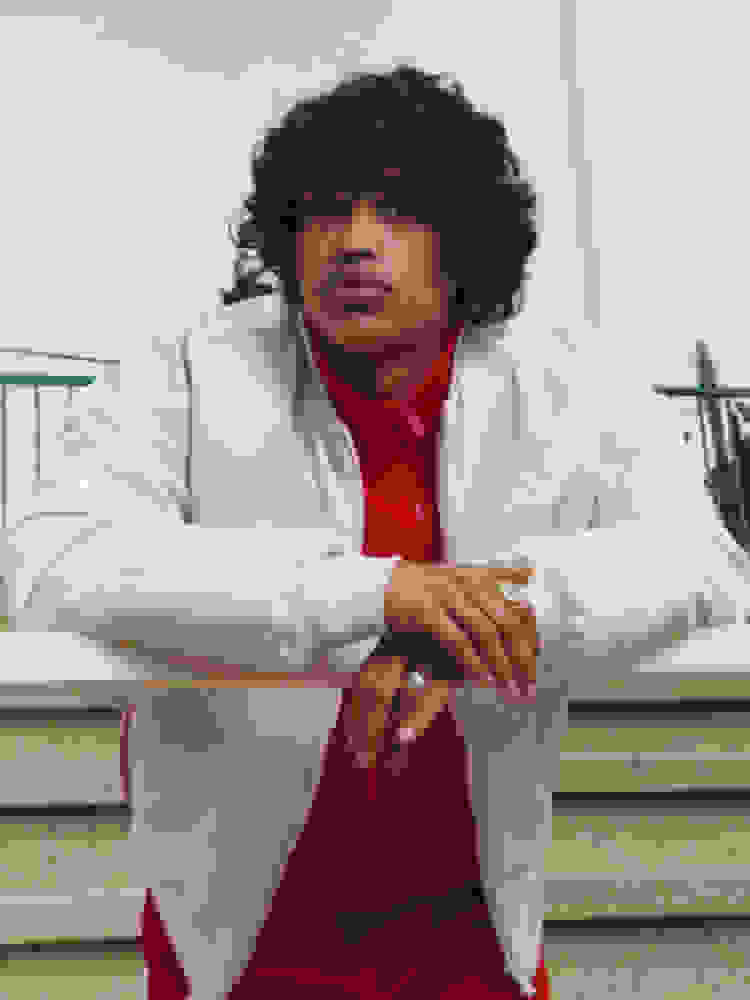 A person with curly dark hair wearing a white jacket and red shirt is sitting on a wooden bench against a backdrop of a clear sky.