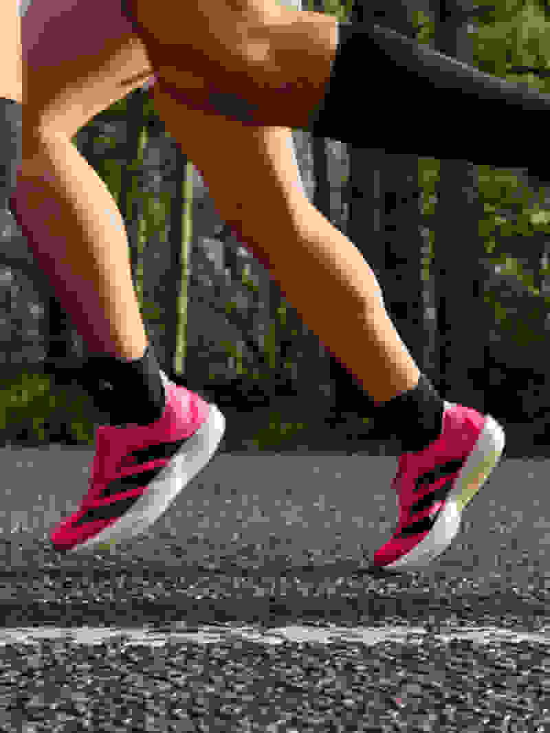 The image shows a person's lower body and legs, wearing bright pink athletic shoes, running on a paved path surrounded by a forested area.