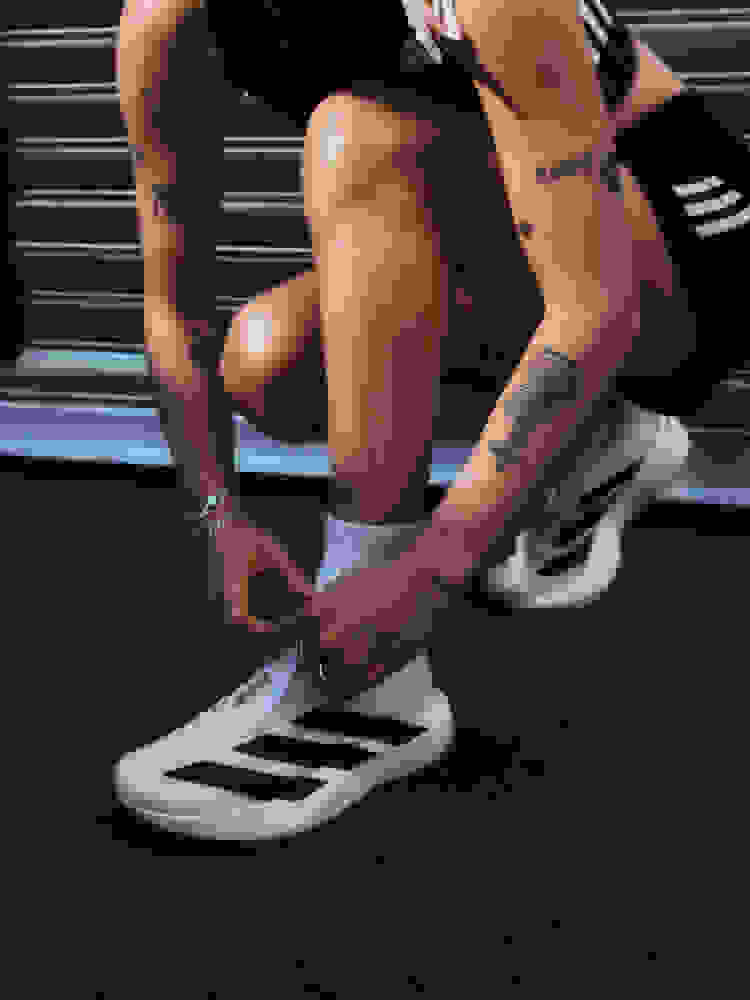 L'immagine mostra la parte inferiore del corpo e delle gambe di una persona, con tatuaggi visibili sulle braccia. Indossano scarpe da atletica bianche e sembrano essere in un ambiente scuro e ombreggiato.