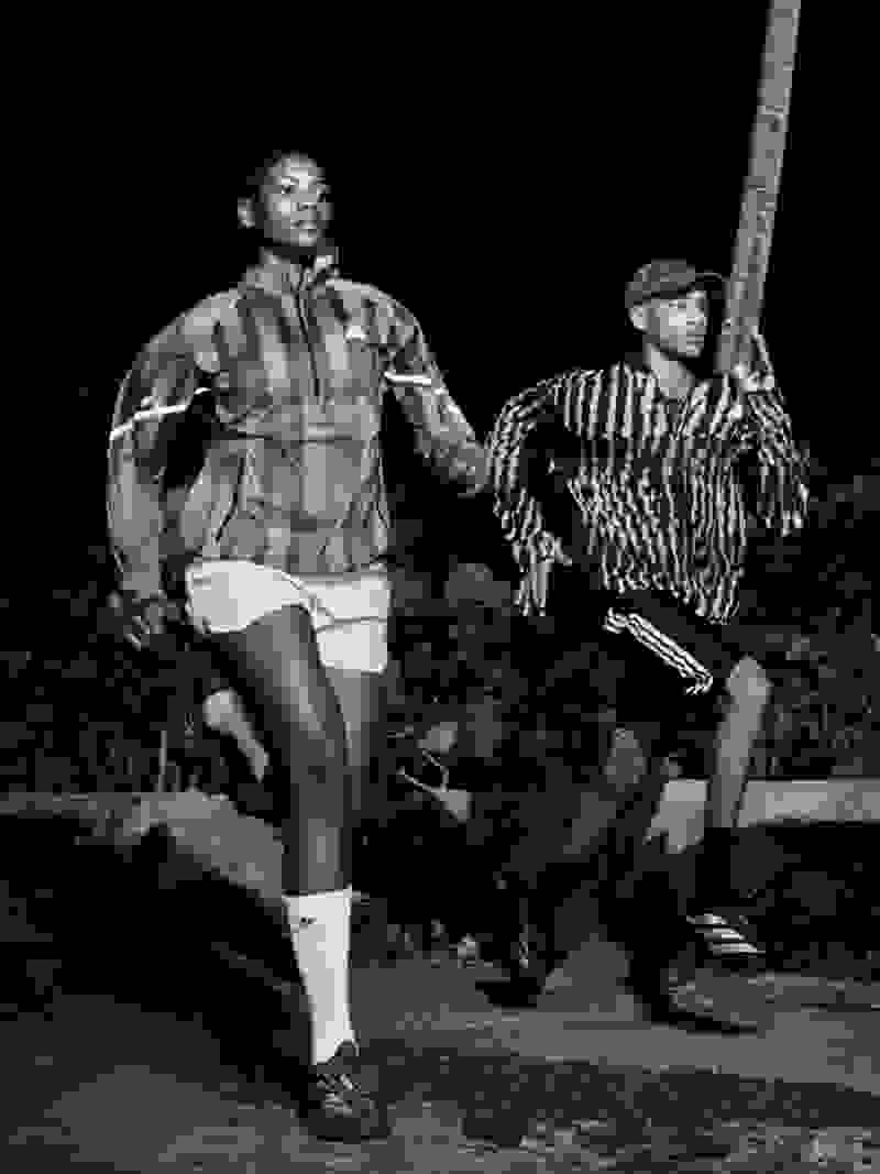 Two individuals, one wearing a plaid shirt and the other a striped shirt, are captured in a black and white photograph, standing in a natural setting with foliage visible in the background.