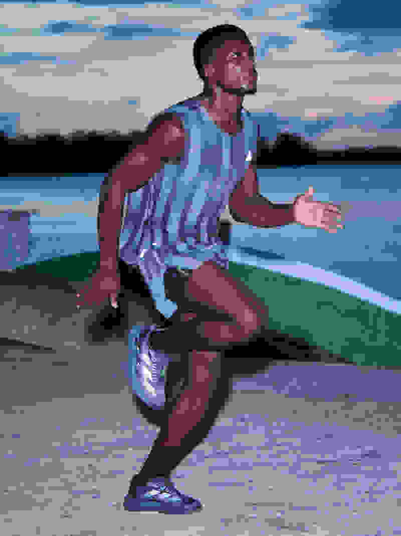 A young man in a striped athletic tank top and shorts is sitting on a dock, with a lake and sunset sky visible in the background.