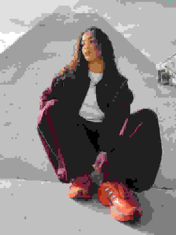 Zeminde bordo palto ve kırmızı sneaker giyen kıvırcık saçlı genç bir kadın, arka planda büyük piramit şekilli bir yapıya sahiptir.