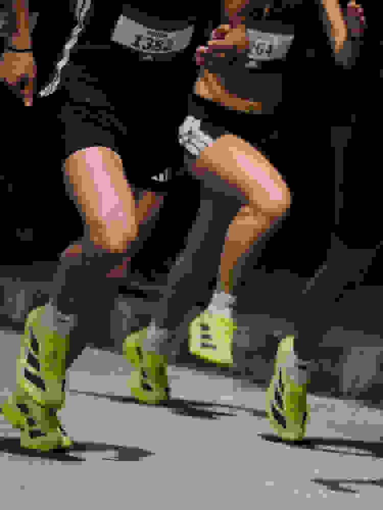 The image shows the lower bodies and legs of runners wearing bright green and black athletic shoes, with race numbers visible, suggesting a running event or competition taking place in a dark environment.