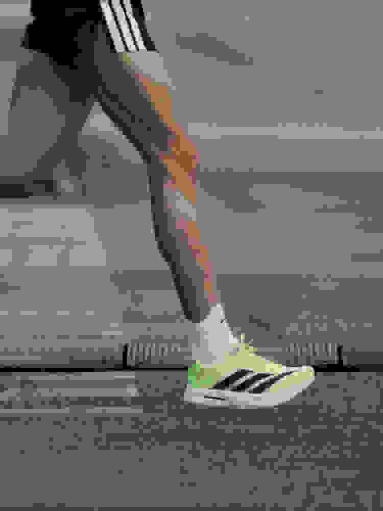 The image shows a person's lower body wearing black athletic shorts and bright green and black running shoes standing on a wooden dock or pier with a body of water in the background.