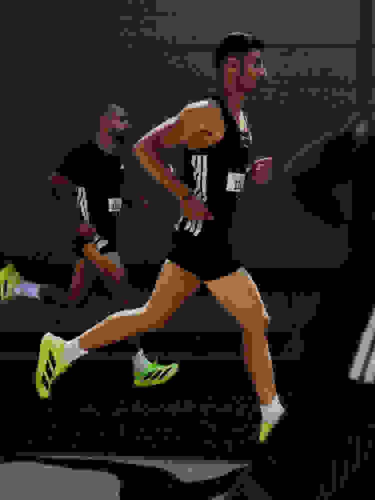 Two male athletes in running gear are competing in a race, with one runner in the foreground and another in the background. The runners are wearing black and white uniforms and bright green running shoes, and the background appears to be a sports facility or track.