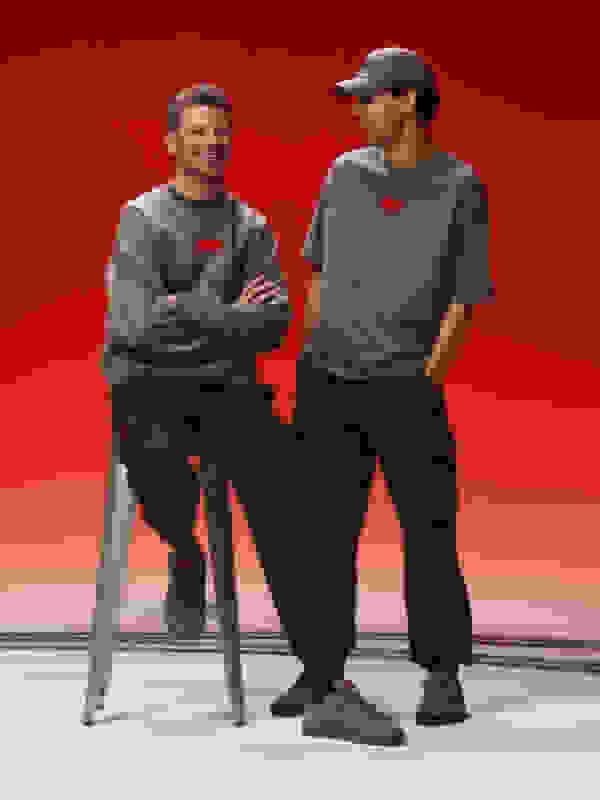 Two individuals wearing casual clothing, including t-shirts and cargo pants, are seated on stools against a vibrant red background.