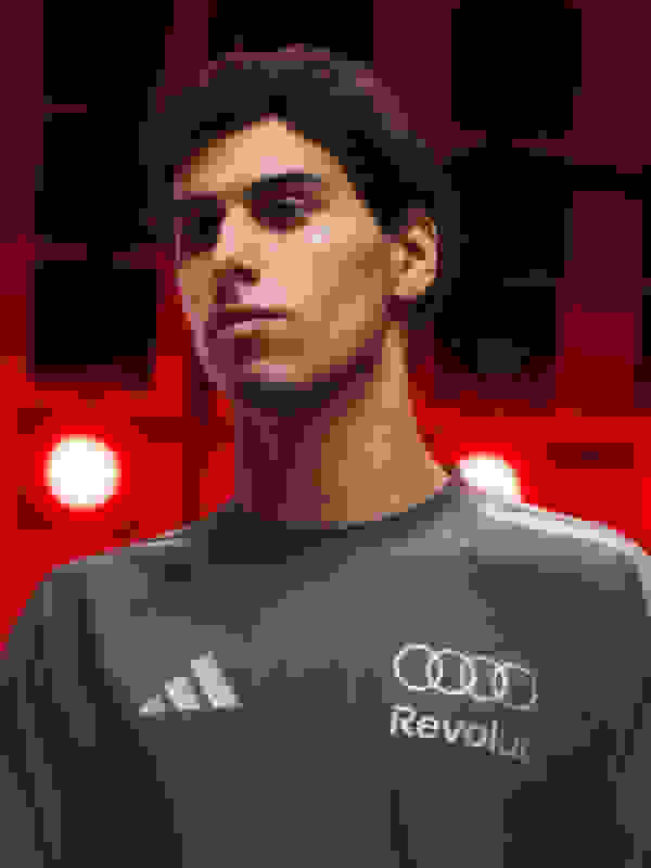 A young man with dark curly hair wearing a gray Adidas shirt with the Audi Revolut logo, standing in front of a red and black background.