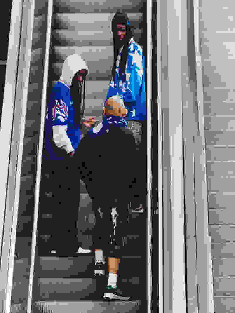 Two individuals, one wearing a blue and white patterned jacket and the other in a black outfit, are standing on an escalator in what appears to be a public transportation setting.