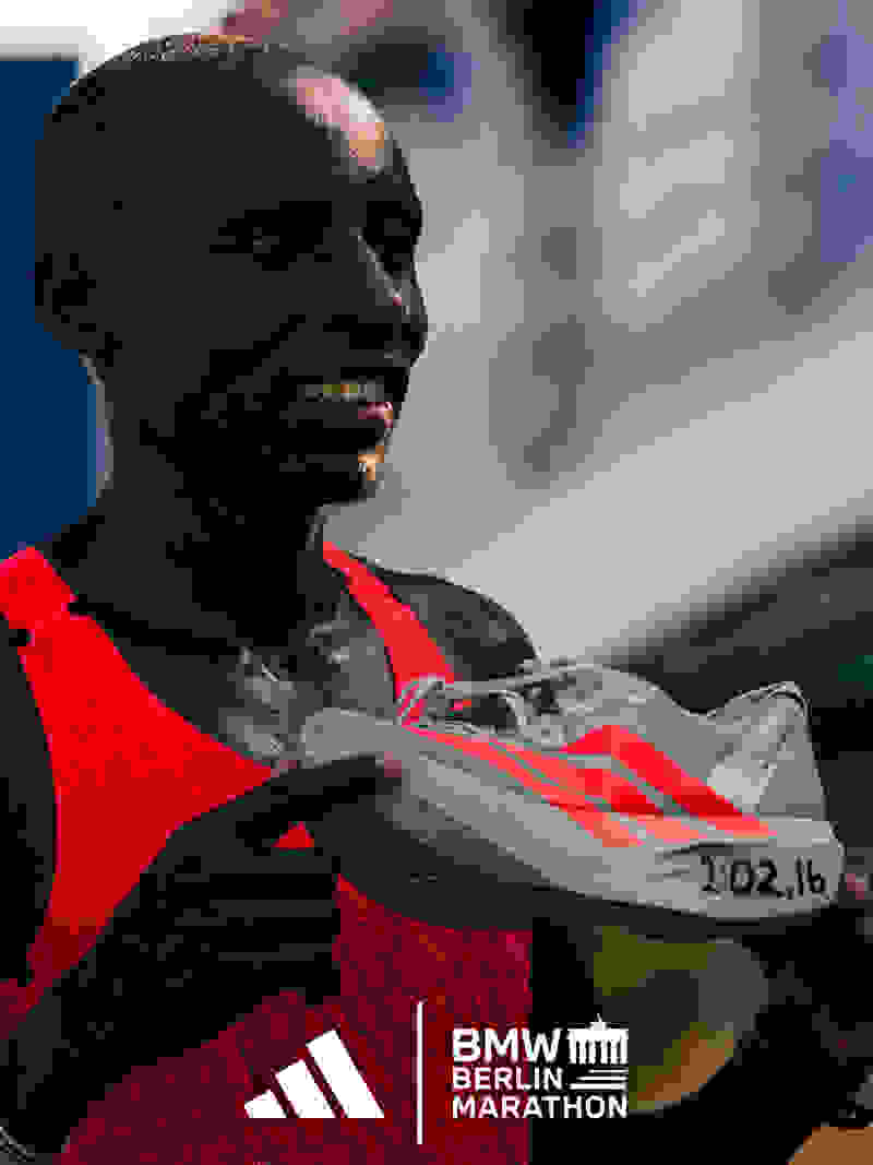A smiling man in a red running jersey holds a pair of running shoes, with the BMW Berlin Marathon logo visible in the background.