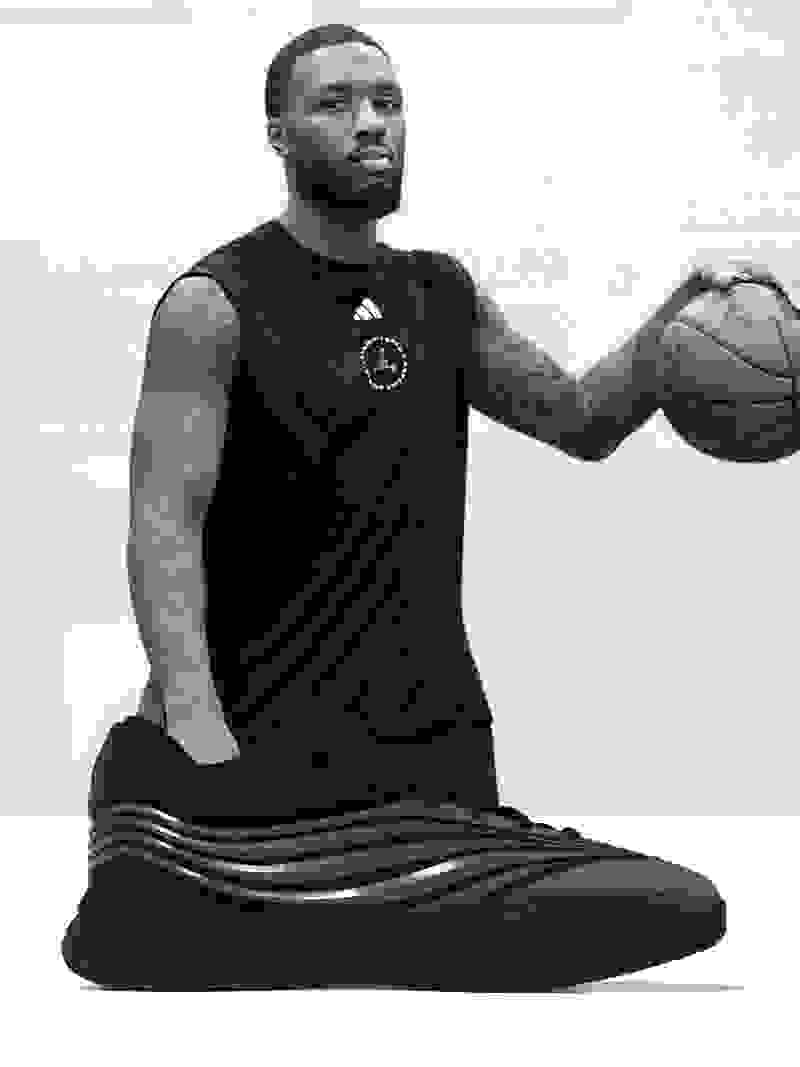 A Black man wearing a black athletic shirt and shorts, sitting on a black exercise mat and holding a basketball.