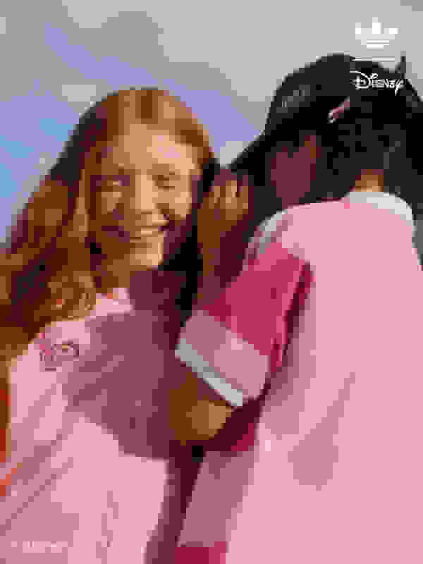 Two young girls, one with red hair and the other with dark hair, are smiling and embracing each other against a backdrop of a cloudy sky.