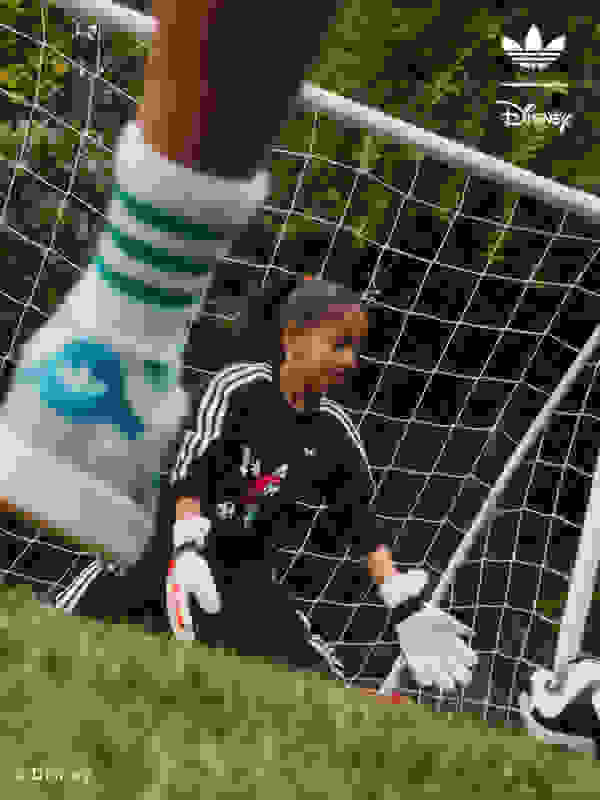 คนหนุ่มสาวในชุดกีฬากำลังนั่งคุกเข่าอยู่บนสนามหญ้าที่ล้อมรอบด้วยรั้วตาข่ายลวด มีโลโก้อาดิดาสและโลโก้ Disney ปรากฏให้เห็นอยู่เบื้องหลัง