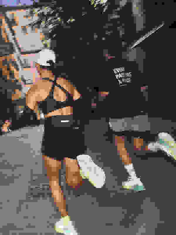 The image shows two people, likely runners, wearing athletic clothing and shoes, moving quickly down a city street at night, with blurred buildings and trees in the background.